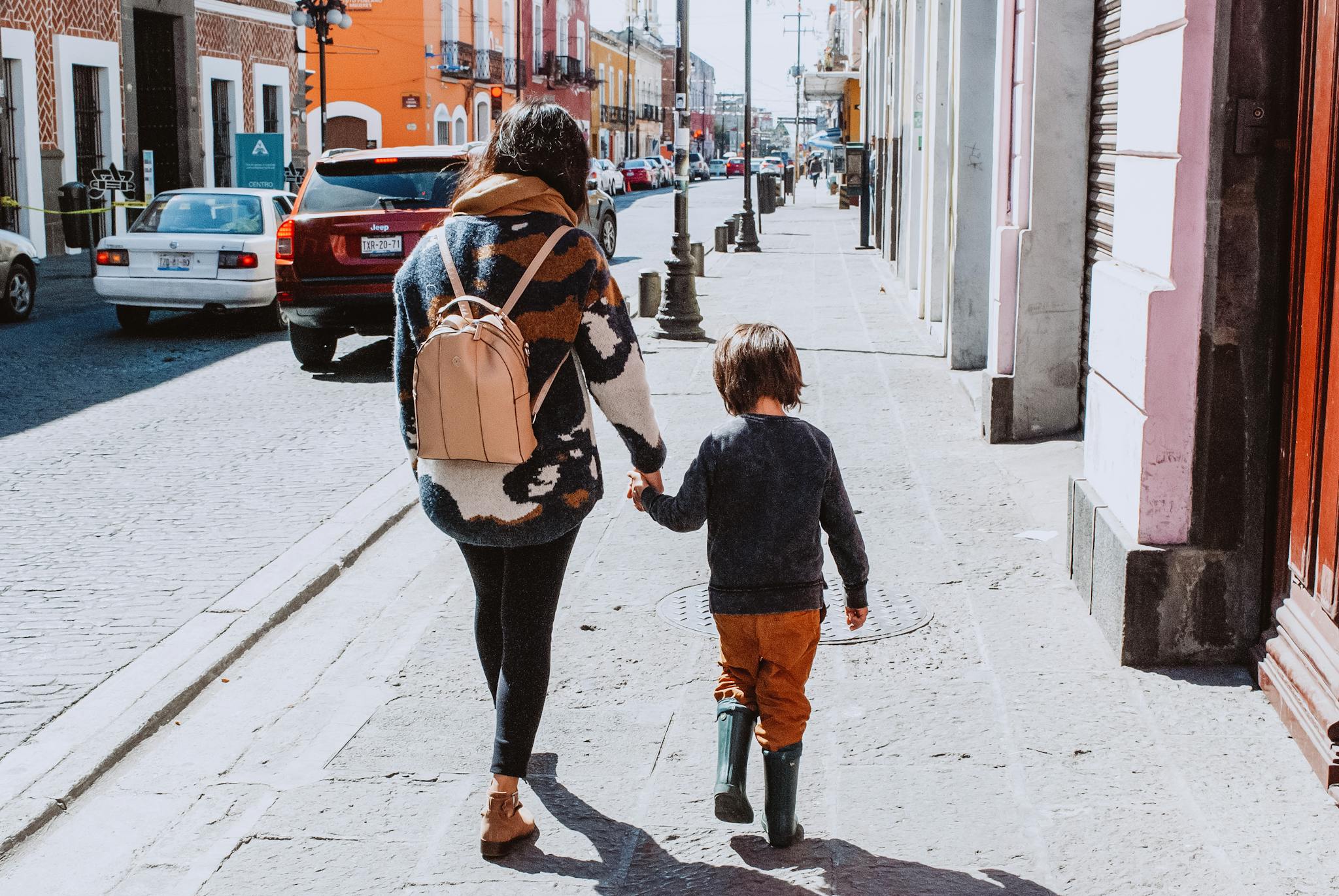 Back view of mother and child holding hands walking on urban sidewalk with colorful buildings.